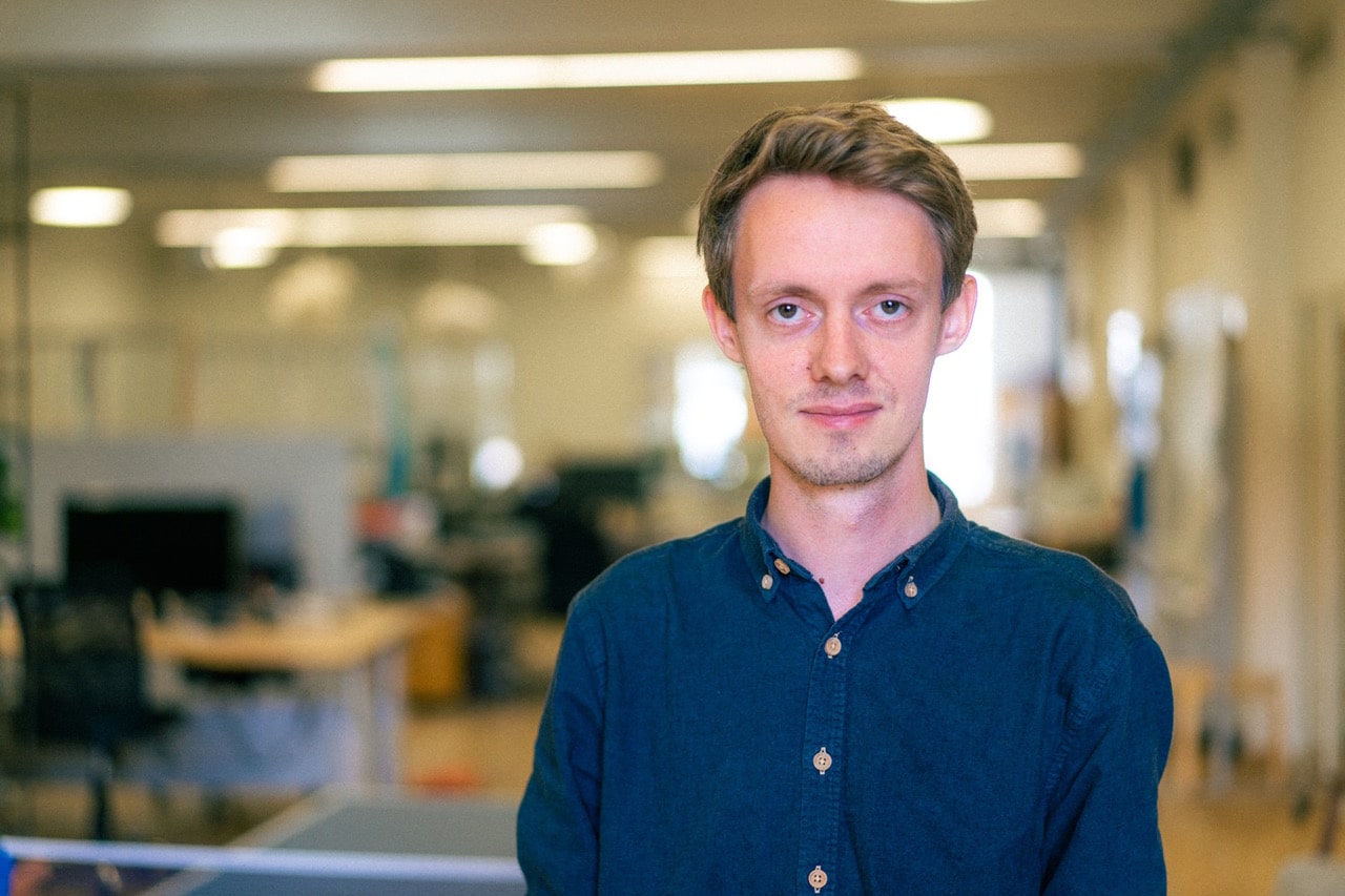 Mads_Skovgaard_raw_1500 En mand med kort hår, iført en blå skjorte, står indendørs i et veloplyst kontorrum med et åbent design. Baggrunden er lidt sløret og fokuserer på ham.